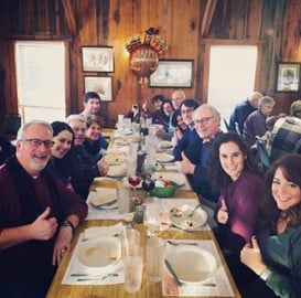 A Corporate group of people seated around a long dining table indoors, smiling and enjoying a shared meal in a warm, rustic setting.