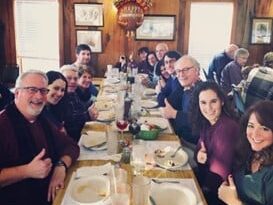 A Corporate group of people seated around a long dining table indoors, smiling and enjoying a shared meal in a warm, rustic setting.