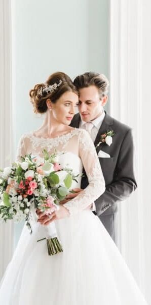 A bride and groom standing together indoors, smiling while holding a bouquet, dressed in wedding attire near a bright window.